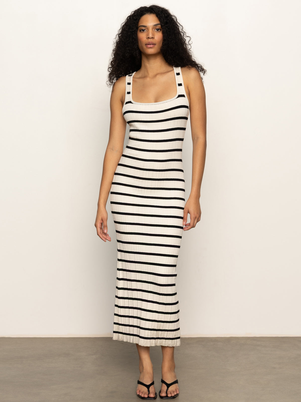 A woman with curly hair wears the Sanctuary Clothing "starlit rib maxi light oat black 3," a sleeveless, fitted, ankle-length striped dress, paired with black sandals, standing against a plain light background.