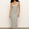 A woman with curly hair wears the Sanctuary Clothing "starlit rib maxi light oat black 3," a sleeveless, fitted, ankle-length striped dress, paired with black sandals, standing against a plain light background.