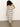 A woman with curly hair wears the Sanctuary Clothing starlit rib maxi light oat black 3, a sleeveless, fitted, black and white striped dress, paired with black sandals. She stands on a concrete floor against a plain white wall.