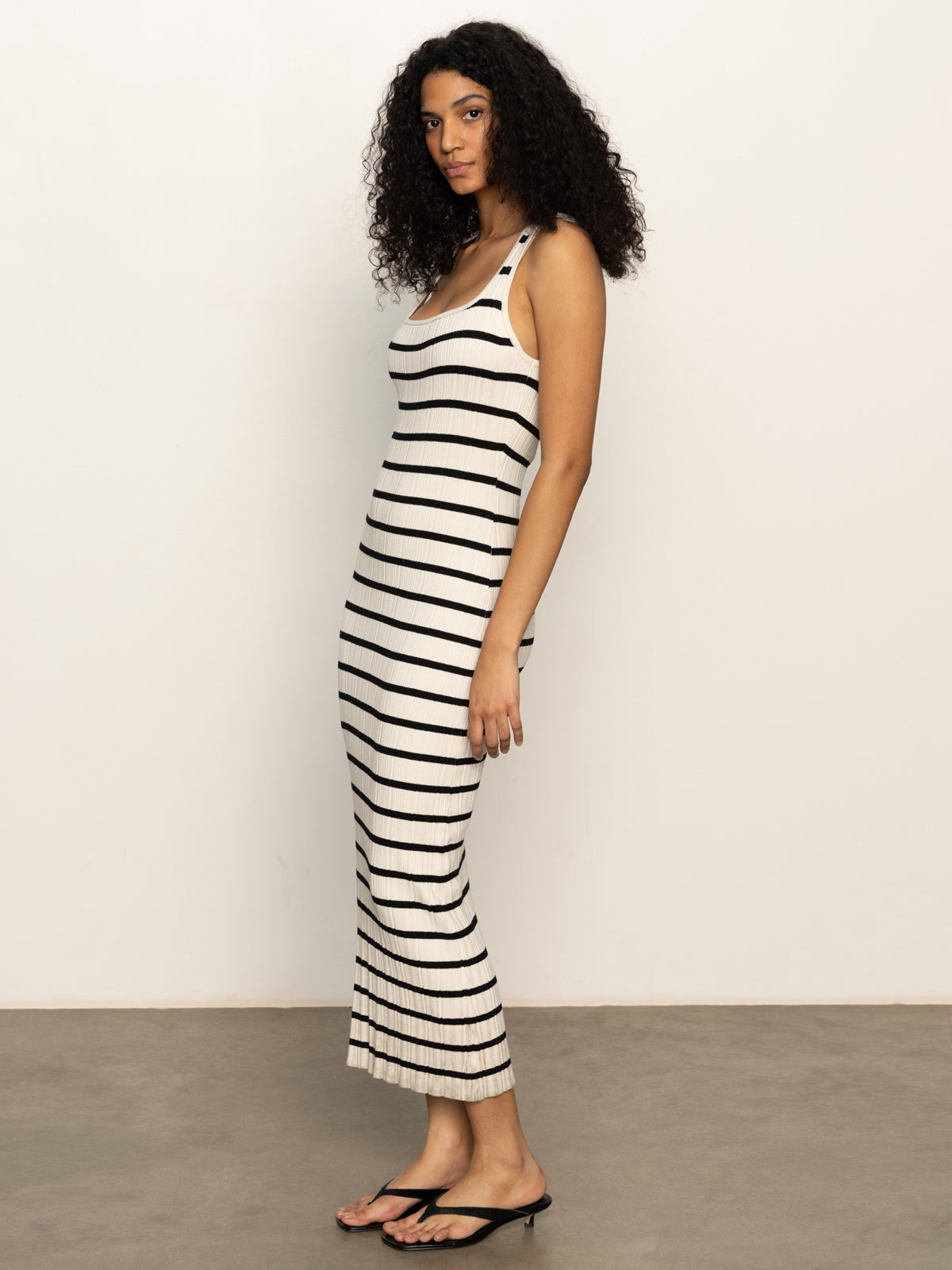 A woman with curly hair stands sideways against a plain background, wearing Sanctuary Clothing's starlit rib maxi light oat black 3, a sleeveless, ankle-length white dress with black stripes and black flip-flops.
