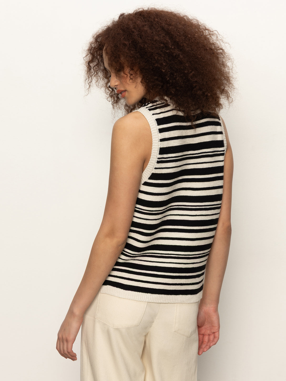 A woman with curly hair, seen from behind against an off-white backdrop, wears the Sanctuary Clothing modern sweater shell in black macrame, paired with light-colored pants.