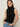 A woman with straight brown hair models the Sanctuary Clothing ribbed funnel neck top in black, paired with light blue jeans. She stands against a plain background with her hands in her pockets, looking directly at the camera.