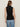 A woman stands with her back to the camera, wearing the Sanctuary Clothing ribbed funnel neck top in black and light blue jeans, set against a plain light-colored background.