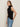 A woman with long brown hair wears the Sanctuary Clothing ribbed funnel neck top in black and light blue jeans, standing against a plain beige background. She looks slightly to the side with a neutral expression.