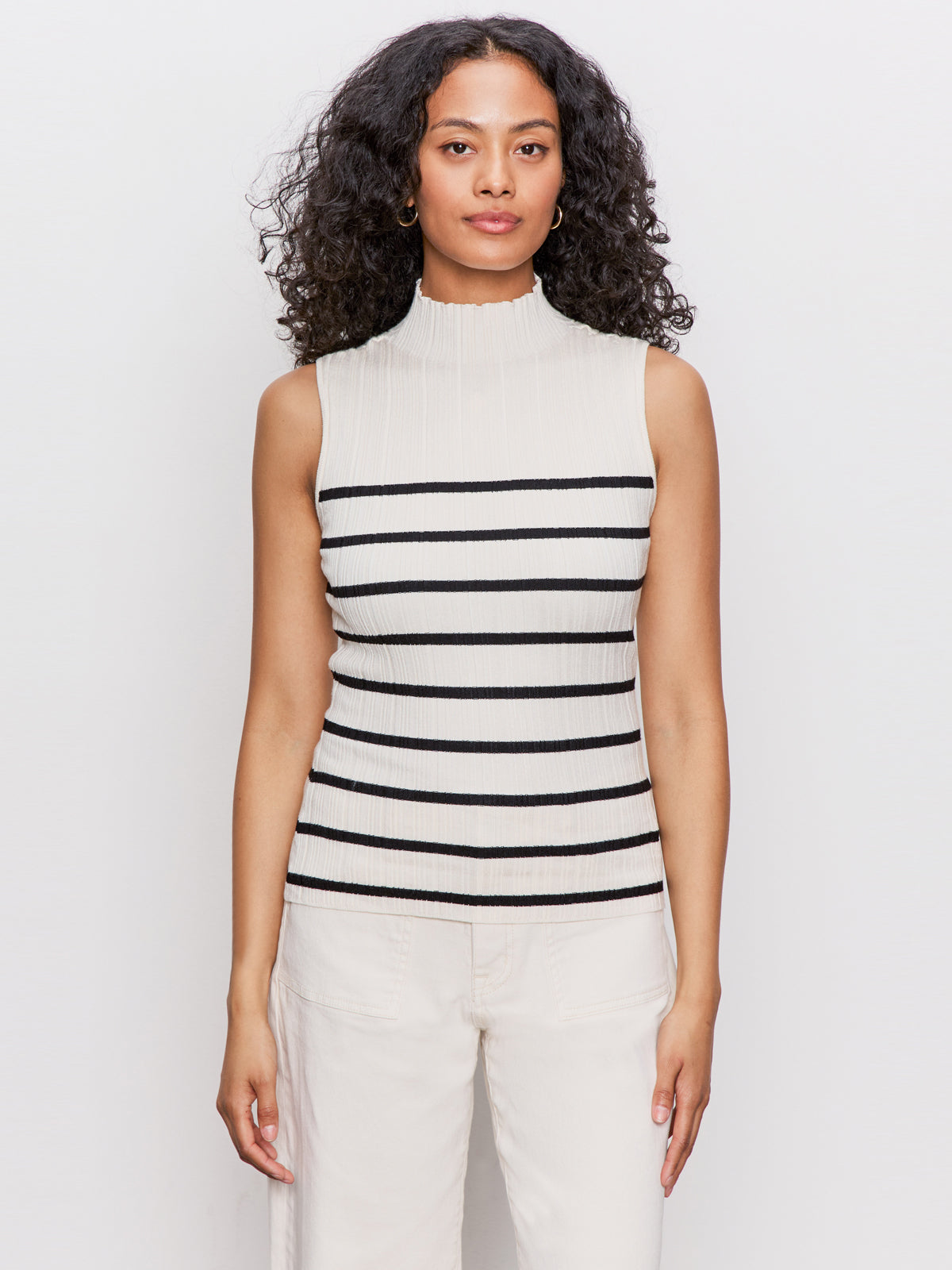A woman with curly hair models the Sanctuary Clothing ribbed funnel neck top in light oat/black, featuring horizontal stripes and a sleeveless design, paired with light pants against a plain white background.