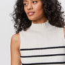A woman with curly dark hair looks to the side, wearing the Sanctuary Clothing ribbed funnel neck top in light oat black 3. The sleeveless top features horizontal black stripes and a mock neckline against a plain white background.