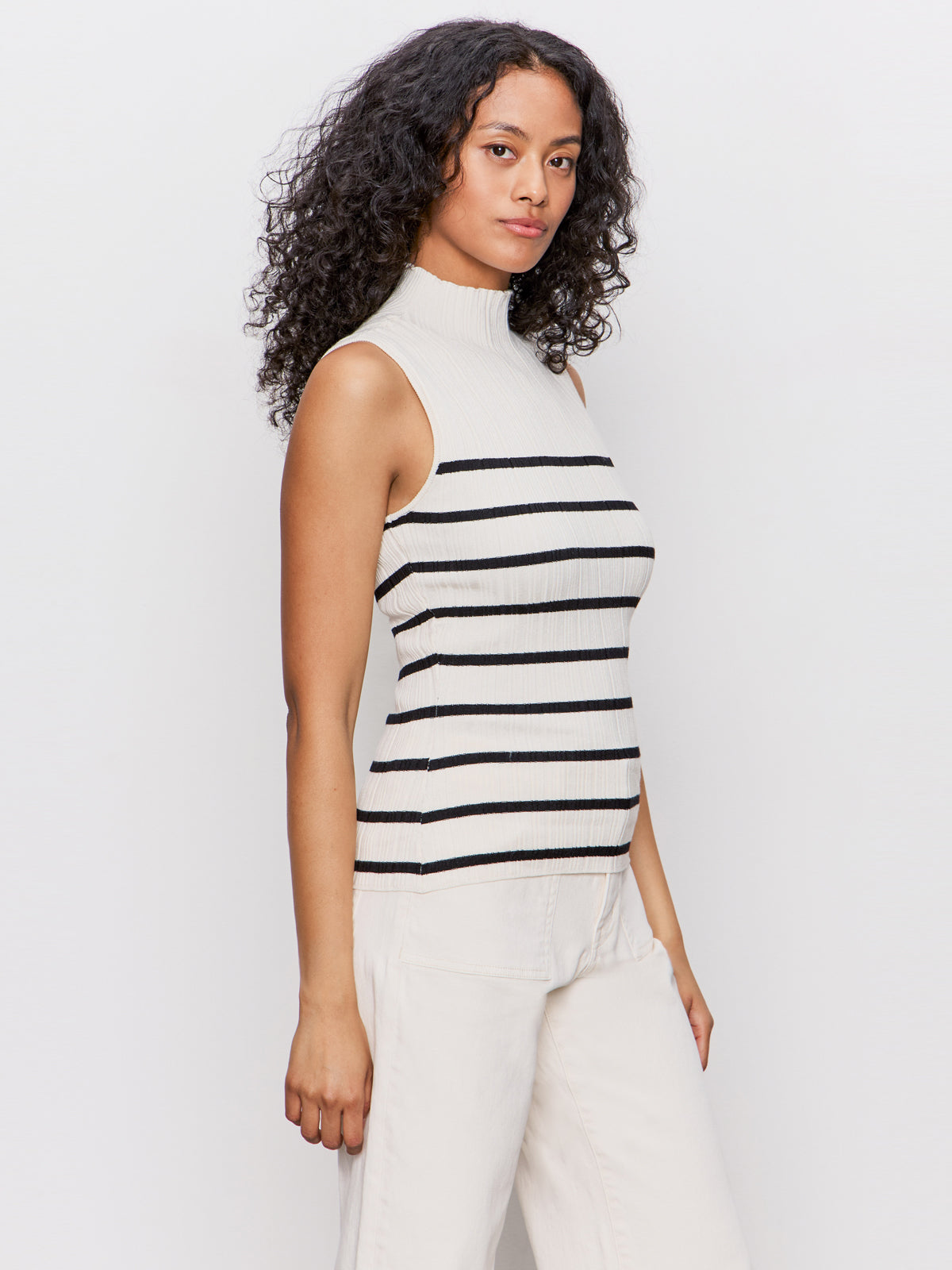 A woman with long curly hair models the Sanctuary Clothing ribbed funnel neck top light oat black 3, a sleeveless high-neck cream top with black stripes, paired with cream pants against a plain white background.