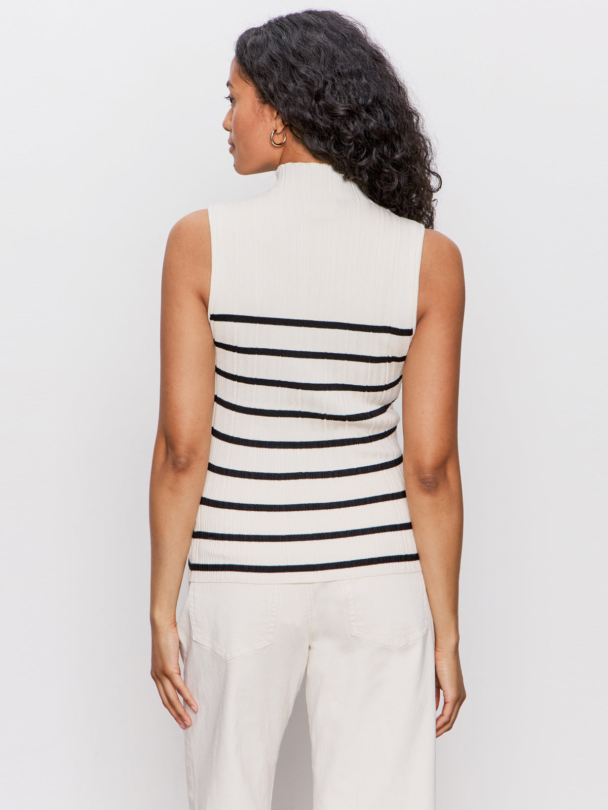 A woman with long curly hair faces away, wearing the Sanctuary Clothing ribbed funnel neck top in light oat black 3 and light-colored pants against a plain white background.