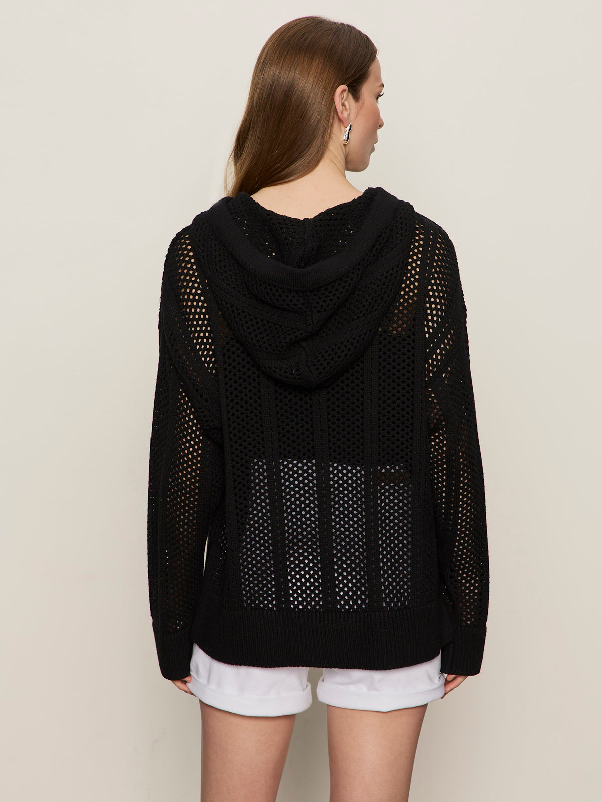 A woman with long brown hair wears the Sanctuary Clothing beach hoodie black, a loosely knit mesh sweater, over white shorts. The see-through hoodie features a relaxed fit and is styled against a plain beige background.