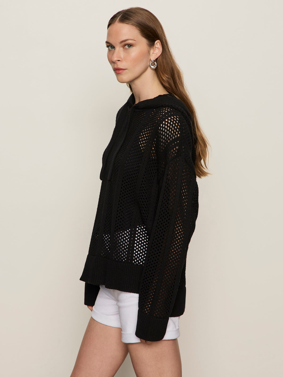 A woman with long brown hair stands sideways against a plain background, wearing Sanctuary Clothing's black beach hoodie over a black top and white shorts. She looks at the camera with a neutral expression.
