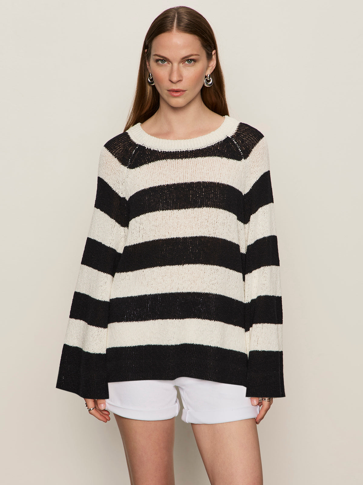 A woman with straight brown hair wears the Sanctuary Clothing textured raglan crew chalk black stripe 6 sweater and white shorts, standing against a plain beige background.