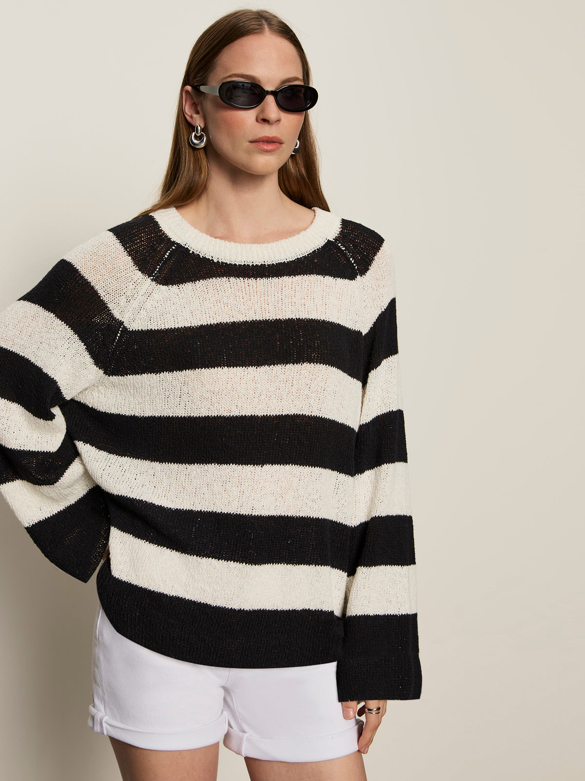 A woman in Sanctuary Clothing's textured raglan crew chalk black stripe 6 poses against a plain background, wearing dark sunglasses, white shorts, hoop earrings, and has straight light brown hair.