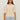 A woman with long brown hair wears the Sanctuary Clothing tape yarn tee macrame (extended sizing), a cream knit short-sleeve sweater, paired with light blue jeans, standing against a plain beige background.