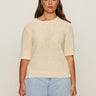 A woman with long brown hair wears the Sanctuary Clothing tape yarn tee macrame (extended sizing), a cream knit short-sleeve sweater, paired with light blue jeans, standing against a plain beige background.