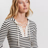 A woman with long blonde hair wears a cream and black striped ribbed top with a lace-up V-neck. She looks slightly to the side against a plain white background.