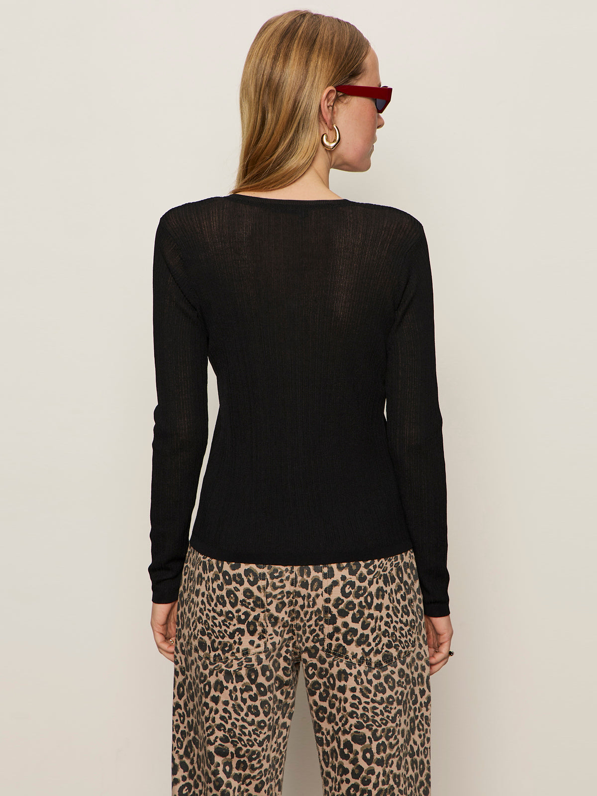 A woman with long blonde hair and red sunglasses wears the Sanctuary Clothing lightweight ribbed cardi in black, leopard print pants, and large hoop earrings, standing with her back to the camera against a plain light background.