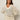 A woman with long brown hair wears the Sanctuary Clothing airy openwork sweater macrame in cream, paired with cream belted pants. She stands against a plain light background, hands on her hips, looking confidently at the camera.