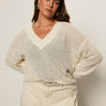 A woman with long brown hair wears the Sanctuary Clothing airy openwork sweater macrame in cream, paired with cream belted pants. She stands against a plain light background, hands on her hips, looking confidently at the camera.