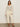 A woman with long brown hair and gold earrings poses confidently in the Sanctuary Clothing airy openwork sweater macrame extended sizing against a plain background, styled with off-white wide-leg pants, a belt, and black heeled sandals.