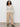 A woman with curly hair stands against a plain background, hands in pockets, wearing the Sanctuary Clothing airy openwork sweater macrame, beige wide-leg pants, and black strappy sandals.