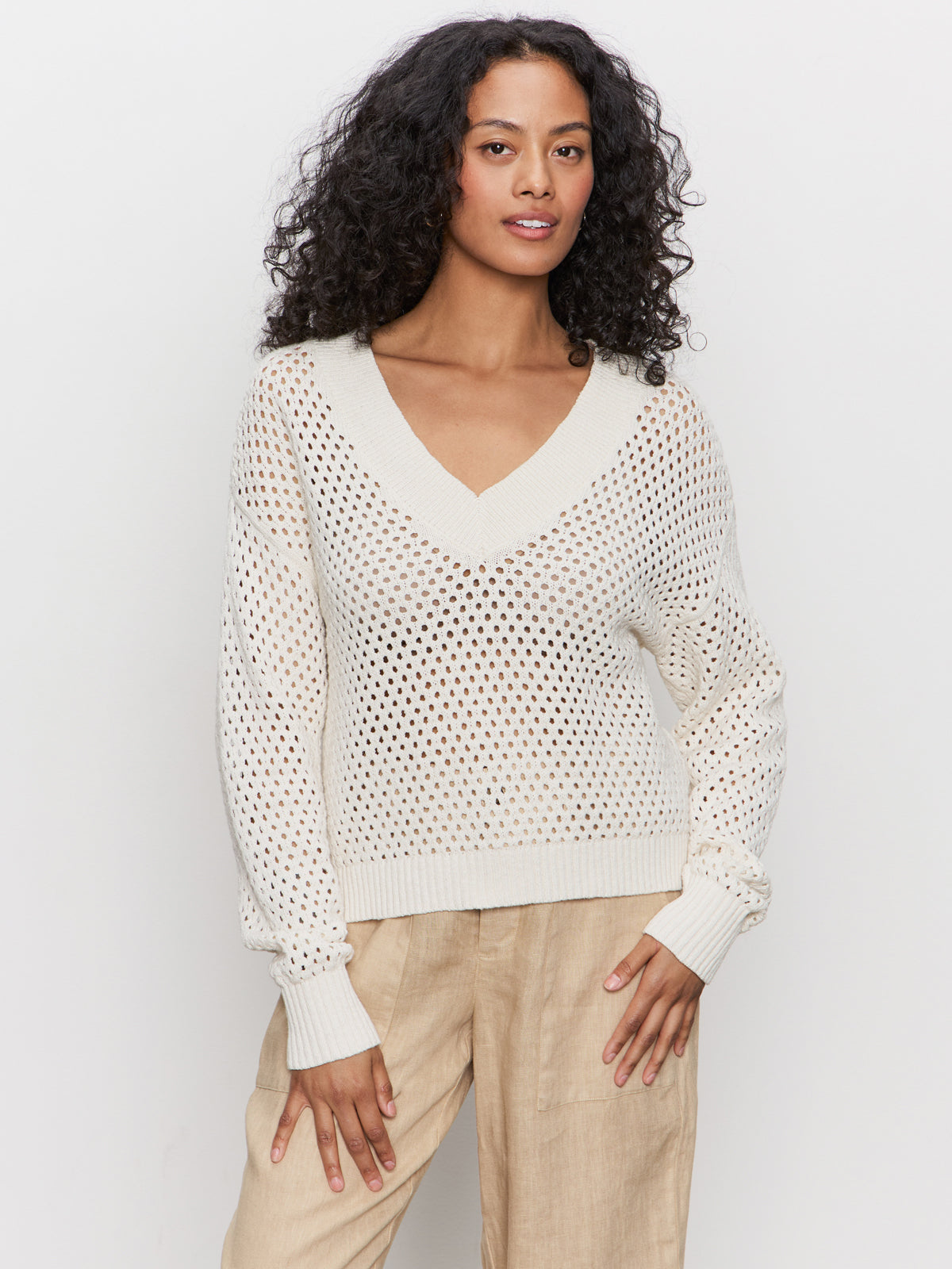 A woman with curly hair wears the Sanctuary Clothing airy openwork sweater macrame in cream and beige pants, standing against a plain white background and looking at the camera with a relaxed expression.