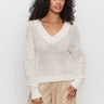 A woman with curly hair wears the Sanctuary Clothing airy openwork sweater macrame in cream and beige pants, standing against a plain white background and looking at the camera with a relaxed expression.
