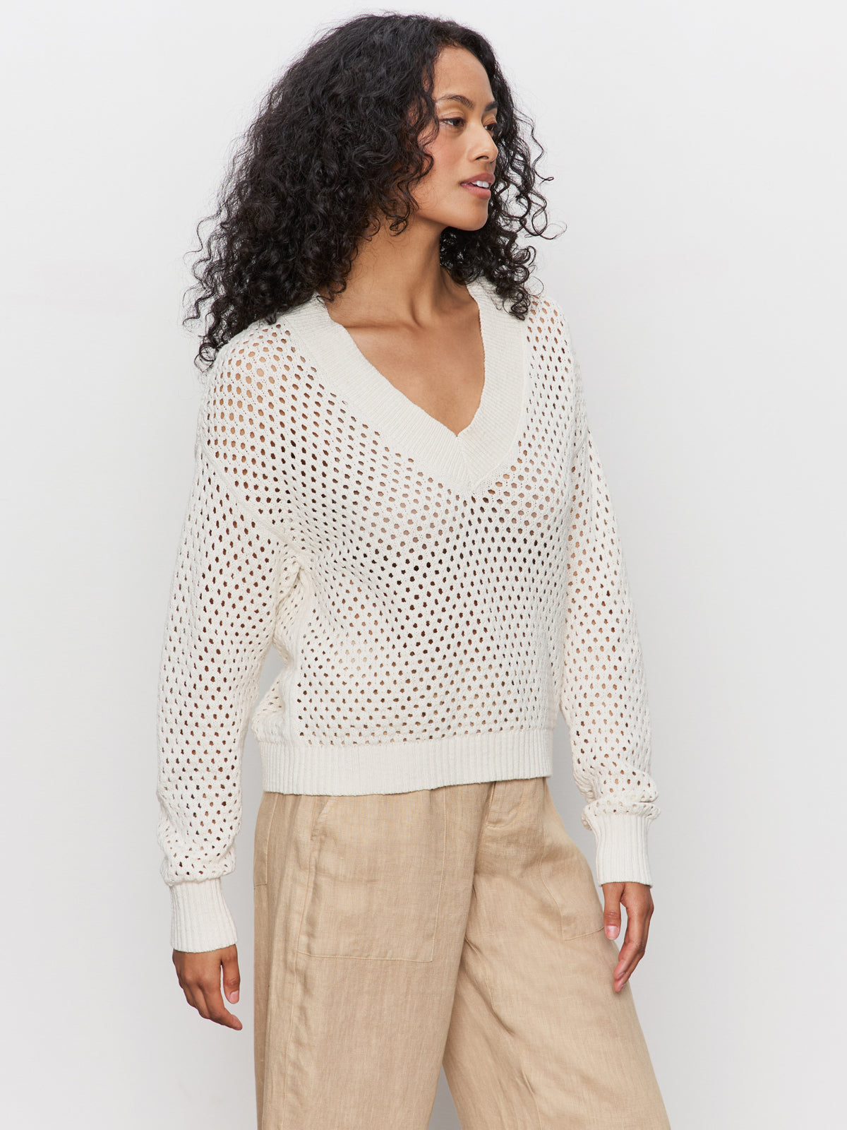 A woman with curly hair wears the Sanctuary Clothing airy openwork sweater macrame in cream over light beige wide-leg pants, standing against a plain white background.