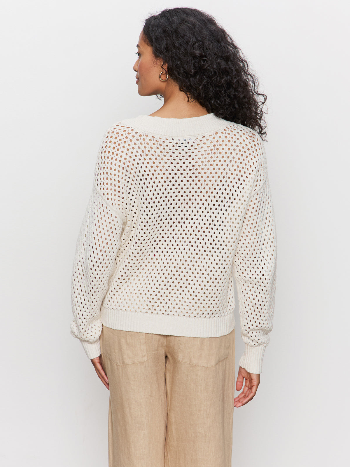 A woman with curly hair wears the Sanctuary Clothing airy openwork sweater macrame in cream, paired with light beige pants, standing with her back to the camera against a plain white background.