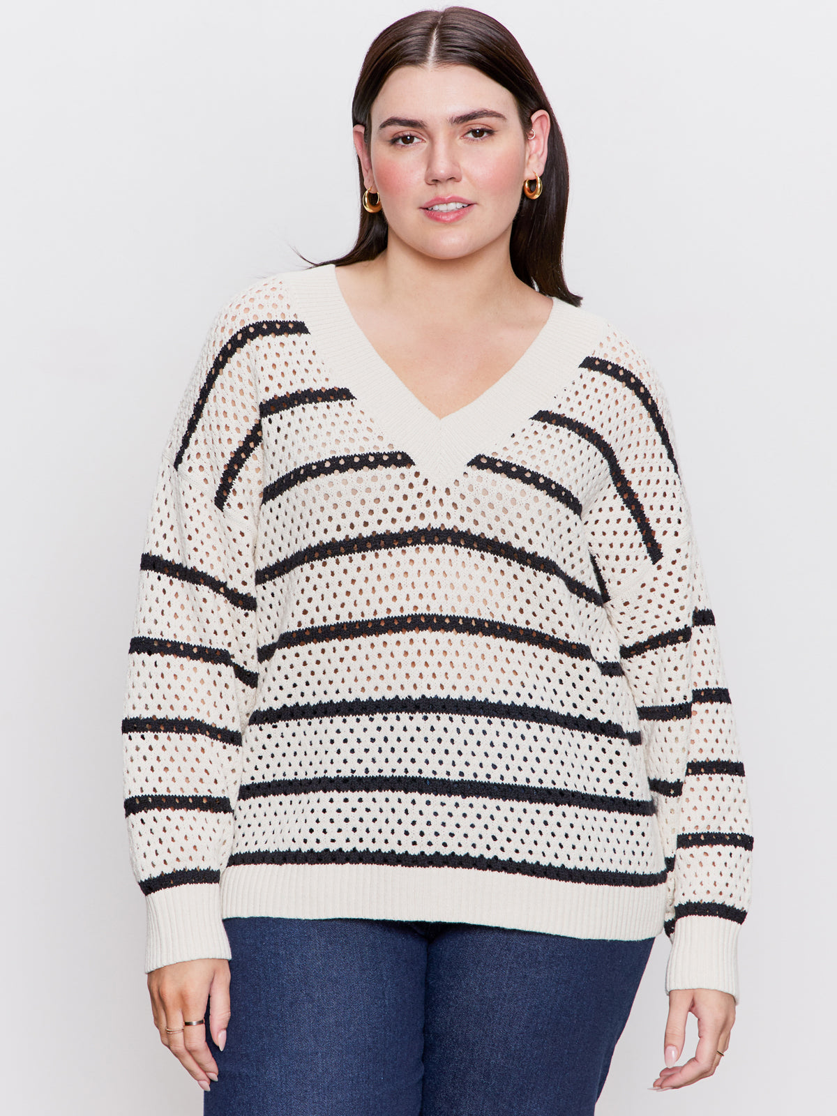 A woman with straight dark hair wears the Sanctuary Clothing airy openwork sweater in chalk black stripe (extended sizing), styled with blue jeans, standing against a plain light background.