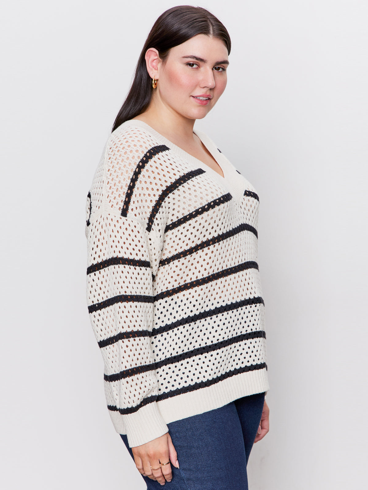 A woman with long dark hair models the Sanctuary Clothing airy openwork sweater in chalk black stripe (extended sizing), paired with blue jeans. She stands sideways, smiling against a plain light backdrop.