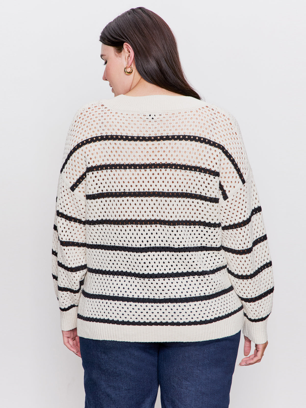 A woman with long dark hair and gold hoop earrings wears the Sanctuary Clothing airy openwork sweater chalk black stripe (extended sizing) and blue jeans, standing with her back to the camera against a plain light background.