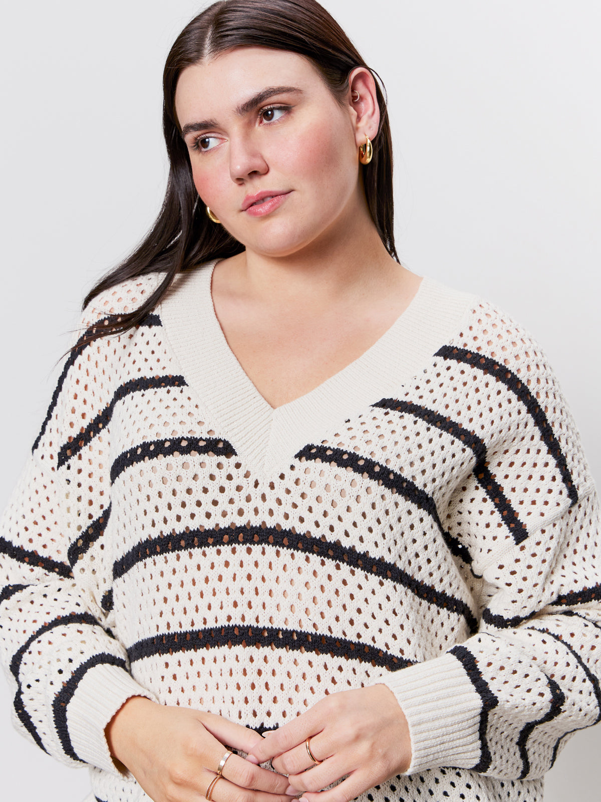 A woman with straight blonde hair wears a cream sweater with black horizontal stripes and light blue jeans. She has one hand in her pocket and wears hoop earrings, standing against a plain light background.