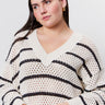 A woman with straight blonde hair wears a cream sweater with black horizontal stripes and light blue jeans. She has one hand in her pocket and wears hoop earrings, standing against a plain light background.