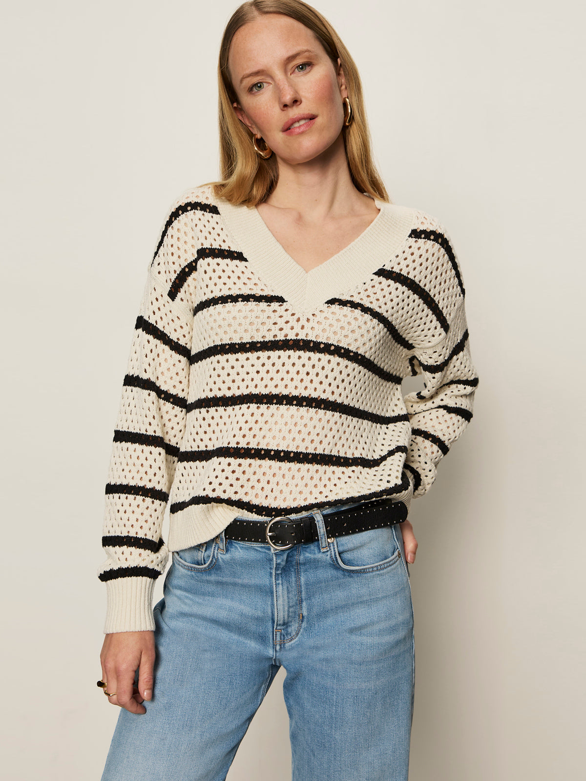 A woman with straight blonde hair wears a cream sweater with black horizontal stripes and light blue jeans. She has one hand in her pocket and wears hoop earrings, standing against a plain light background.