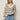 A woman with straight blonde hair wears a cream sweater with black horizontal stripes and light blue jeans. She has one hand in her pocket and wears hoop earrings, standing against a plain light background.