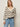 A woman with straight blonde hair wears a cream sweater with black horizontal stripes and light blue jeans. She has one hand in her pocket and wears hoop earrings, standing against a plain light background.