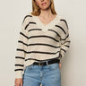 A woman with straight blonde hair wears a cream sweater with black horizontal stripes and light blue jeans. She has one hand in her pocket and wears hoop earrings, standing against a plain light background.