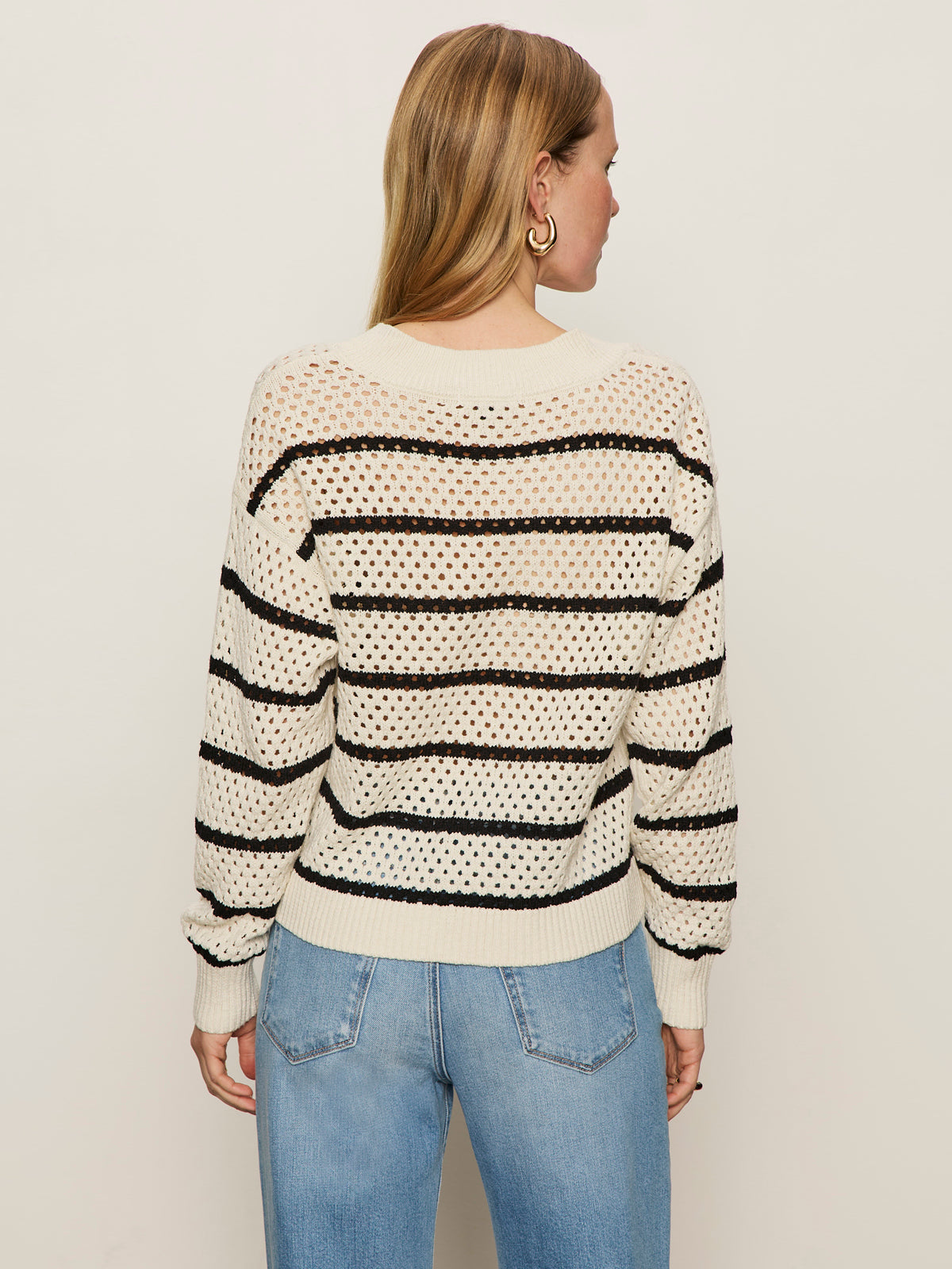 A woman with long, light brown hair wears the Sanctuary Clothing airy openwork sweater in chalk black stripe and blue jeans, standing with her back to the camera against a plain, light background.