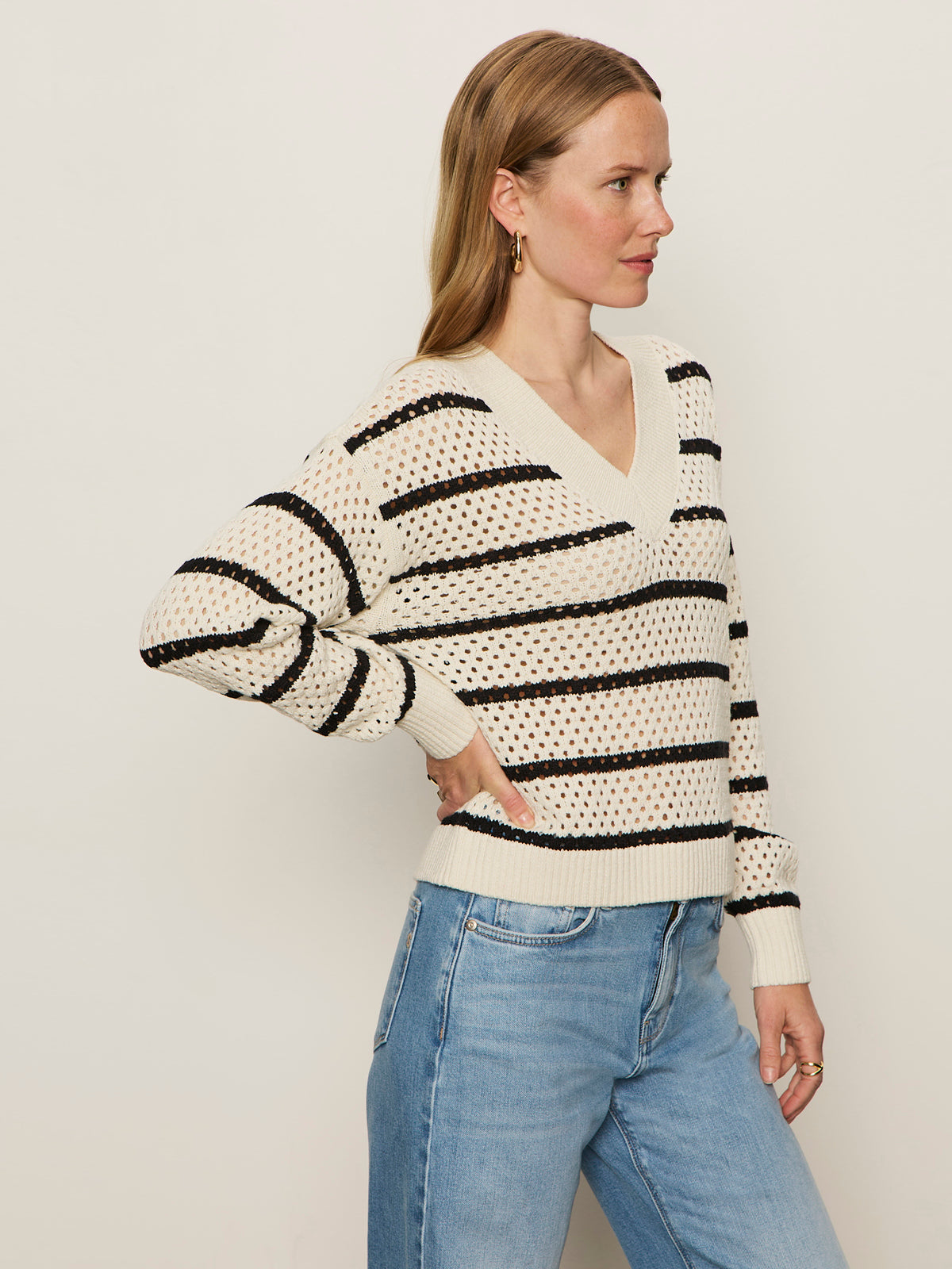A woman with light brown hair and a neutral expression stands sideways against a plain background, wearing Sanctuary Clothing's airy openwork sweater in chalk black stripe, paired with light blue jeans and gold hoop earrings.