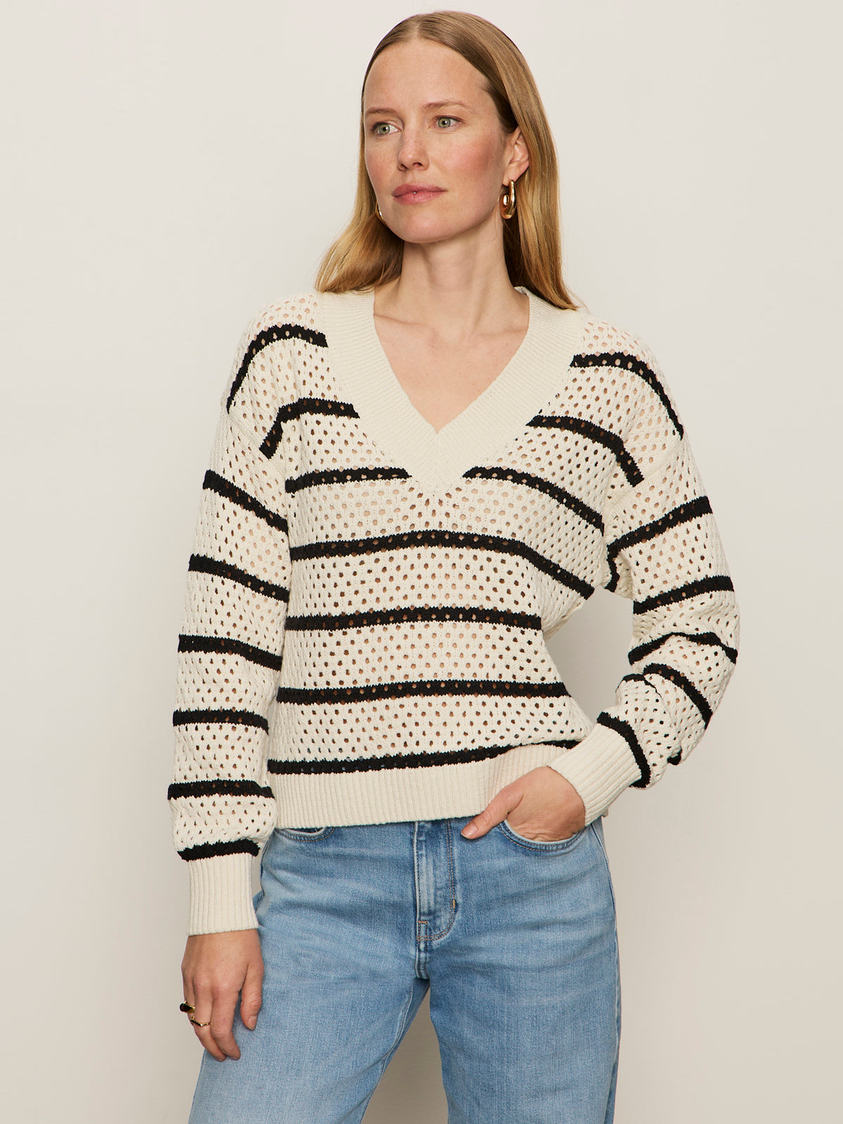 A woman with long blonde hair models the Sanctuary Clothing airy openwork sweater in chalk black stripe, paired with light blue jeans, standing against a plain light background.