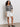 A woman wears the Sanctuary Clothing city summer cardi black macrame multi stripe, paired with white shorts and black woven flats, standing against a plain white wall on a concrete floor.