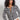 A woman with curly hair models the Sanctuary Clothing city summer cardi in black macrame multi stripe, paired with white pants, standing against a plain light background with her hands in her pockets.
