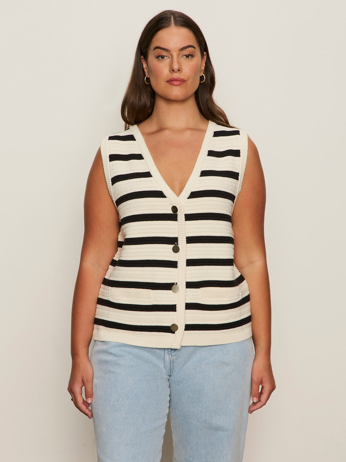 A woman with long brown hair wears the Sanctuary Clothing textured sweater vest macrame black stripe 4, a sleeveless V-neck knit top with black and white stripes, paired with light blue jeans against a plain light background.