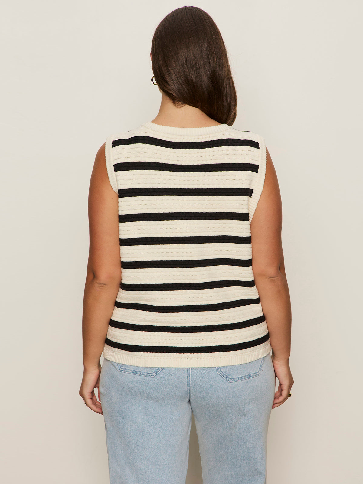 A person with long brown hair faces away, modeling the Sanctuary Clothing textured sweater vest macrame black stripe 4 with light blue jeans against an off-white background.