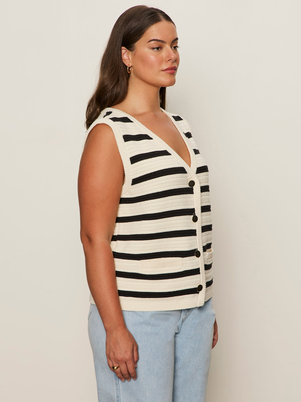 A woman with long brown hair stands in profile, wearing the Sanctuary Clothing textured sweater vest macrame black stripe 4—a sleeveless, v-neck cream vest with bold black stripes and buttons—paired with light blue jeans against a plain background.