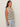 A woman with long brown hair stands in profile, wearing the Sanctuary Clothing textured sweater vest macrame black stripe 4—a sleeveless, v-neck cream vest with bold black stripes and buttons—paired with light blue jeans against a plain background.