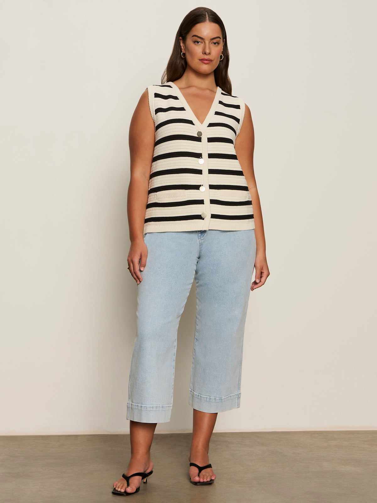 A woman poses confidently in the Sanctuary Clothing textured sweater vest macrame black stripe 4, paired with light blue cropped jeans, black sandals, and hoop earrings against a plain wall.