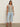 A woman poses confidently in the Sanctuary Clothing textured sweater vest macrame black stripe 4, paired with light blue cropped jeans, black sandals, and hoop earrings against a plain wall.