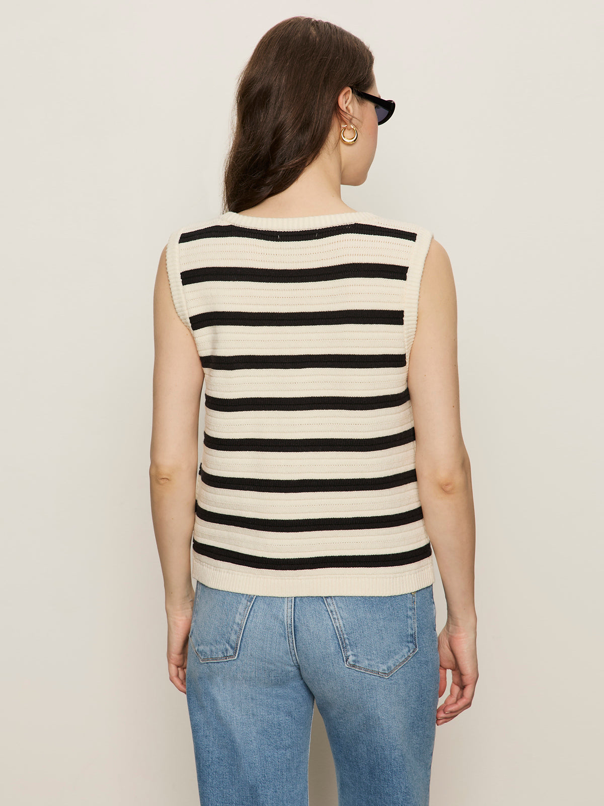 A woman with long brown hair, sunglasses, and blue jeans models the Sanctuary Clothing textured sweater vest macrame black stripe 3, standing with her back to the camera against a plain light background.