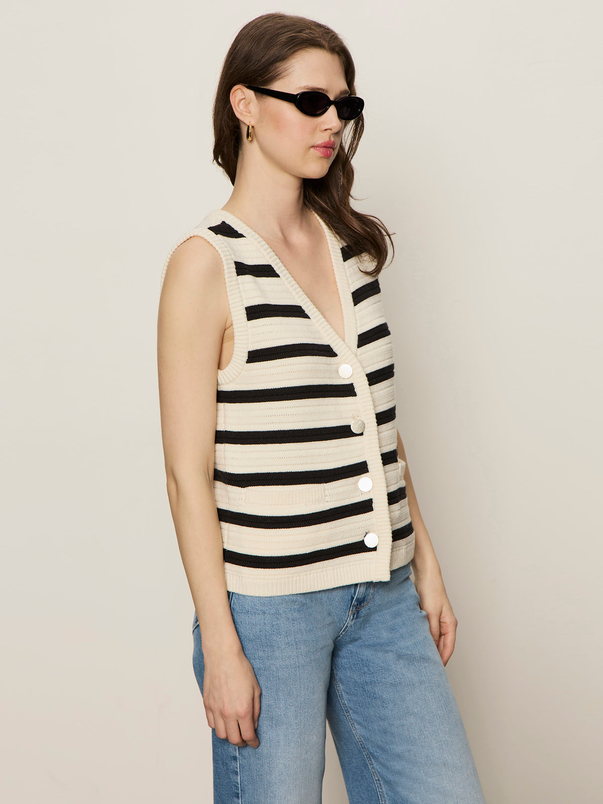 A woman stands in front of a plain light-colored background wearing Sanctuary Clothing's textured sweater vest macrame black stripe 3, sunglasses, and blue jeans.